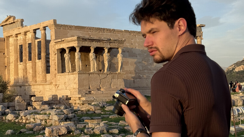Photographer holding a camera while standing in front of ancient Greek temple ruins during golden hour.