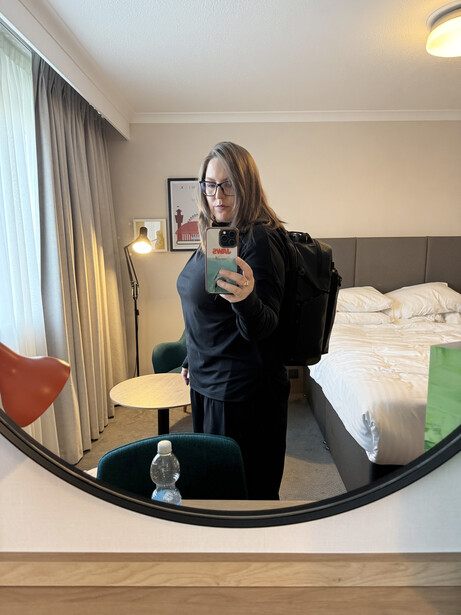 Woman taking a mirror selfie in a hotel room, holding a glass toward the camera.