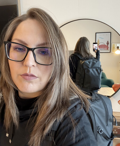 Woman wearing glasses and dark jacket looks directly at camera in indoor setting with mirror visible behind her.