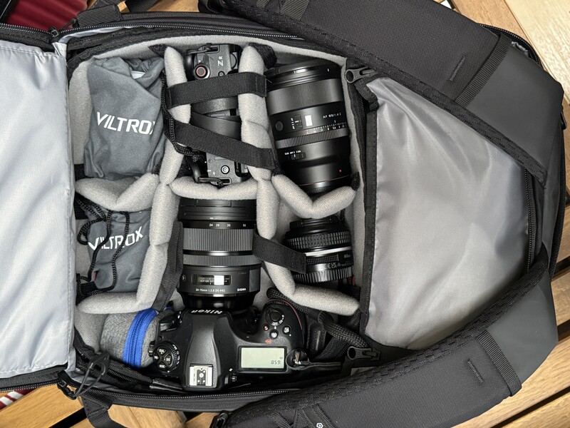Open camera backpack displaying organized photography gear including DSLR body, multiple lenses, and accessories wrapped in protective padding.