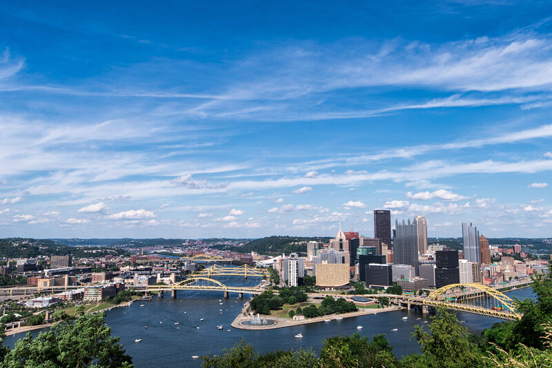 Aerial cityscape with downtown skyline, three bridges spanning a river, and forested hills under blue sky.
