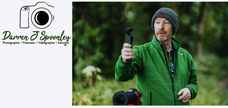 Videographer in green jacket holding gimbal stabilizer outdoors with blurred forest background.