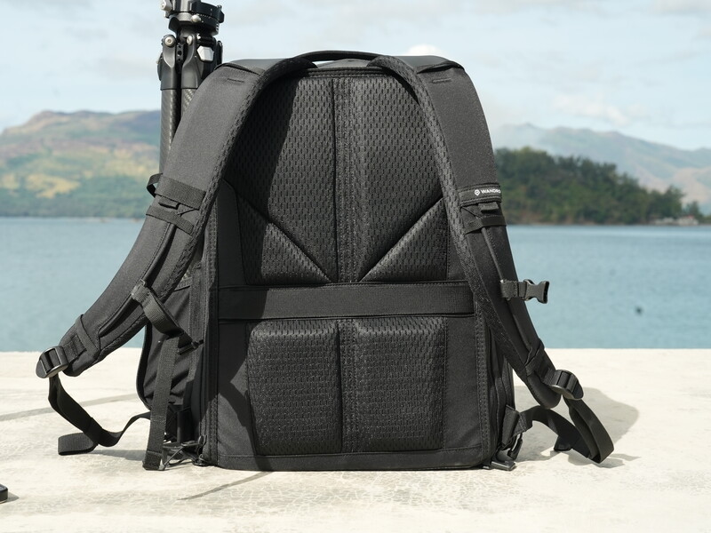 Black camera backpack with mesh ventilation panels displayed on sandy beach with water and mountains in background.