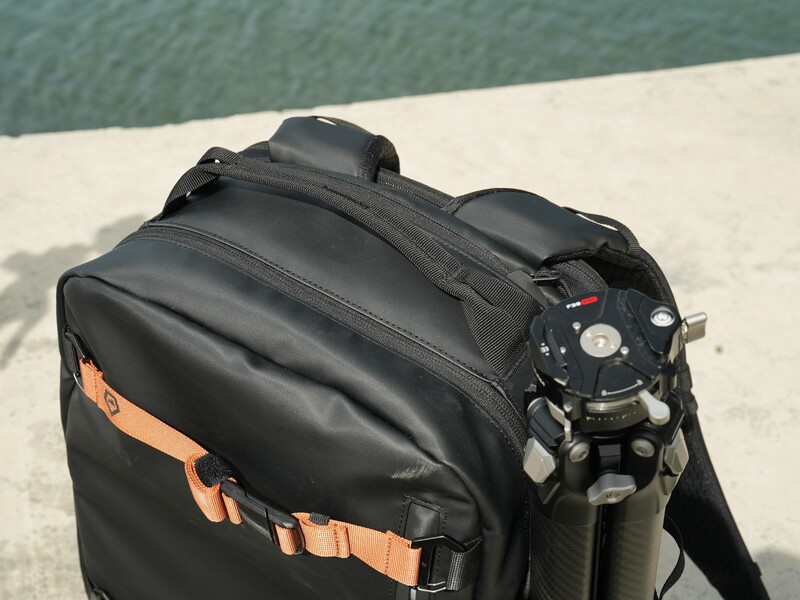 Black camera backpack with orange accent straps displayed next to a mirrorless camera body on a concrete surface near water.