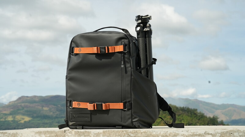 Dark gray camera backpack with orange accents and attached tripod against mountainous landscape.