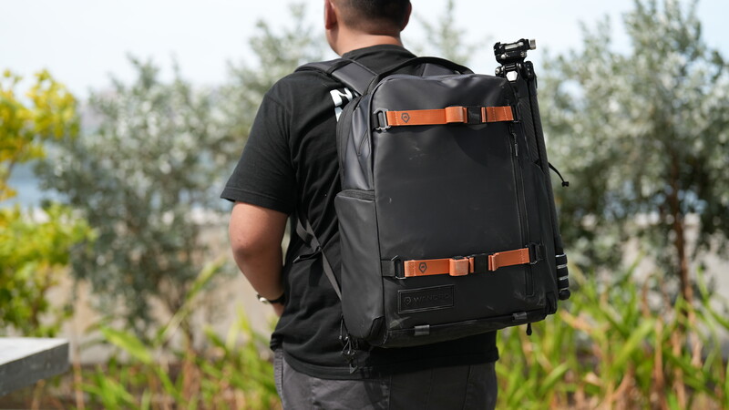 Photographer wearing black travel backpack with orange straps outdoors among trees.
