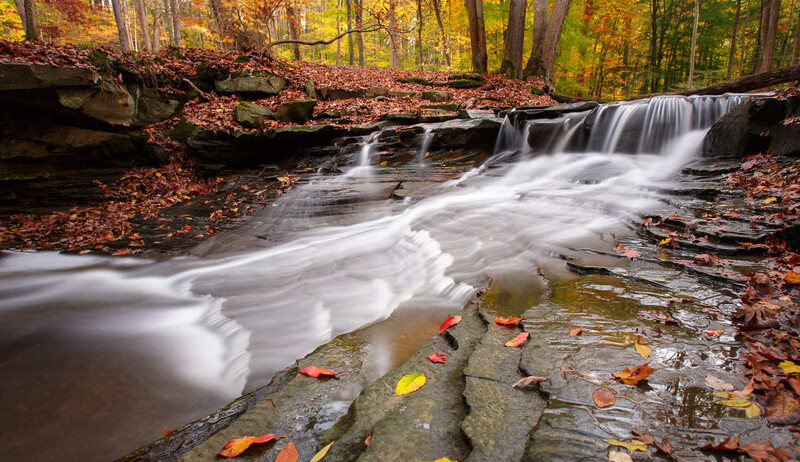 Cascading waterfall flowing over layered rock formations surrounded by autumn foliage and fallen leaves.