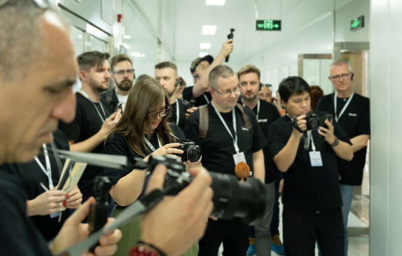 Group of photographers in black polo shirts with lanyards holding cameras and recording equipment in a modern hallway.