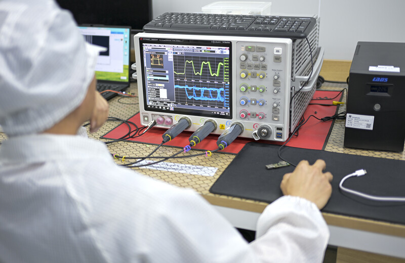 Technician in white lab coat operates an oscilloscope displaying waveform signals on its screen.