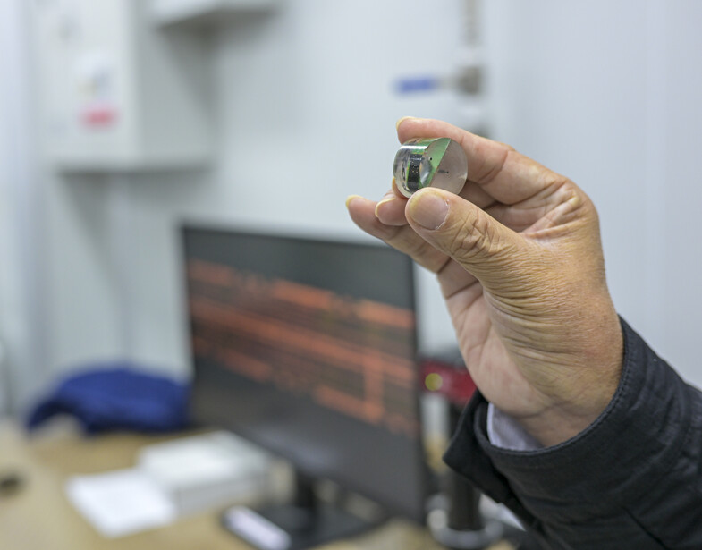 Person holding a small electronic component or microchip in focus, with a blurred computer monitor and office setting in the background.