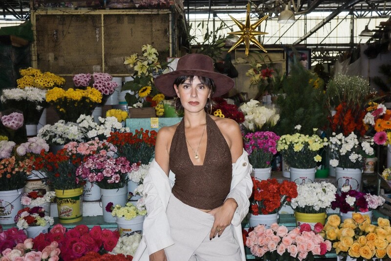 Woman in brown vest and white shirt poses among vibrant cut flowers in a greenhouse setting.