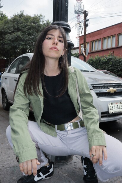 Woman in light green jacket and gray pants crouching near parked vehicles on urban street.