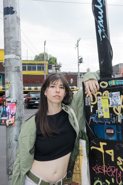 Woman in black crop top and olive jacket posing against urban graffiti wall and pole.