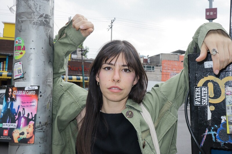 Woman with long dark hair in green jacket against urban street backdrop with utility pole and brick buildings.