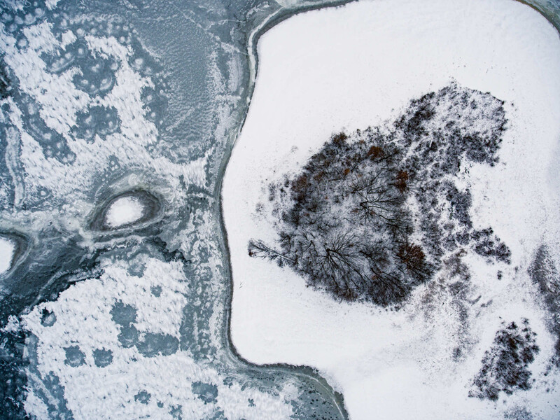 Aerial view of snowy landscape with dark forested island and winding frozen waterway.