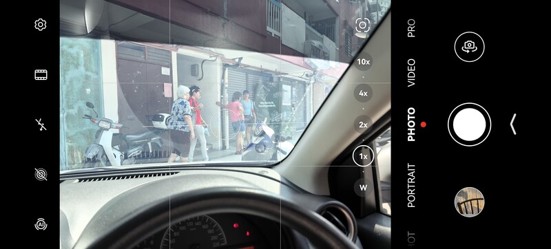 Screenshot of smartphone camera interface showing street scene through windshield with zoom and mode controls visible.