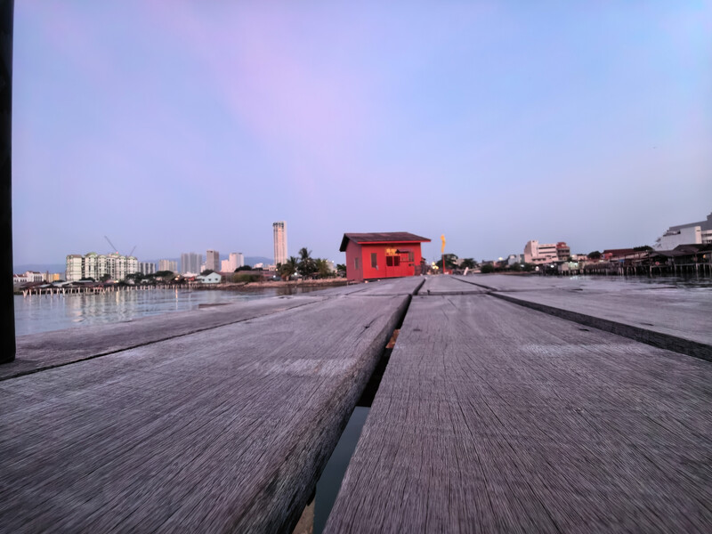 Red wooden structure on a wet wooden boardwalk at dusk, with city skyline and water reflections.