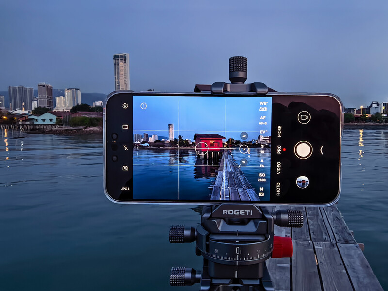 Smartphone mounted on a gimbal capturing waterfront cityscape with red boathouse and wooden pier.