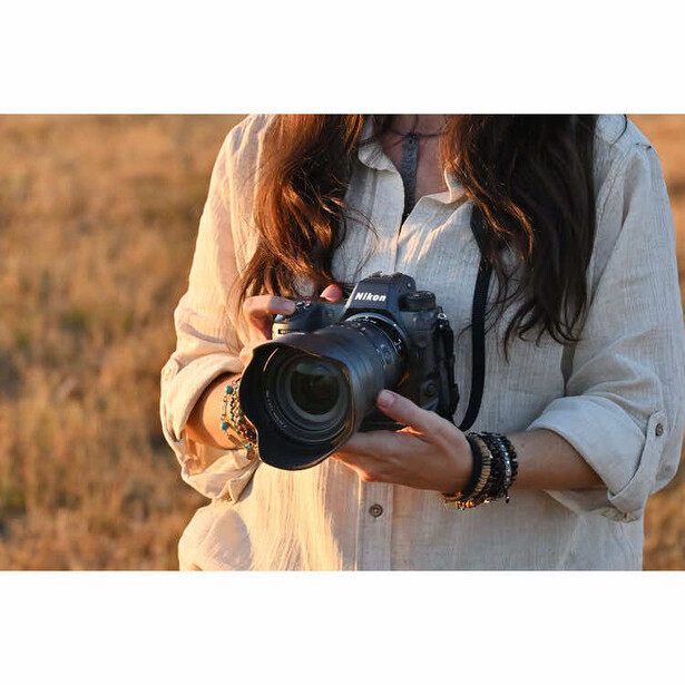 Photographer holding a DSLR camera with telephoto lens, wearing light linen shirt in golden hour sunlight.