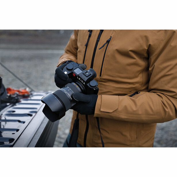 Photographer in brown jacket holding a mirrorless camera with telephoto lens outdoors.