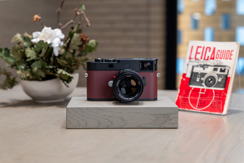 Burgundy Leica M rangefinder camera displayed on concrete block with guide booklet and potted flowers.