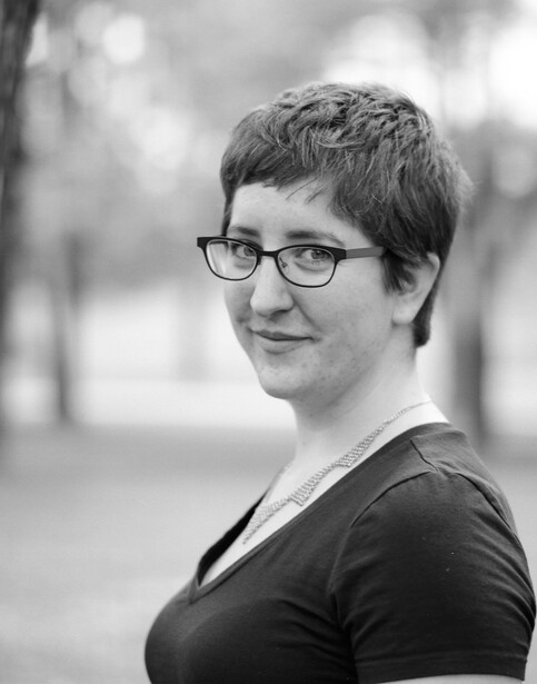 Portrait of a person wearing glasses and dark clothing against a blurred outdoor background.