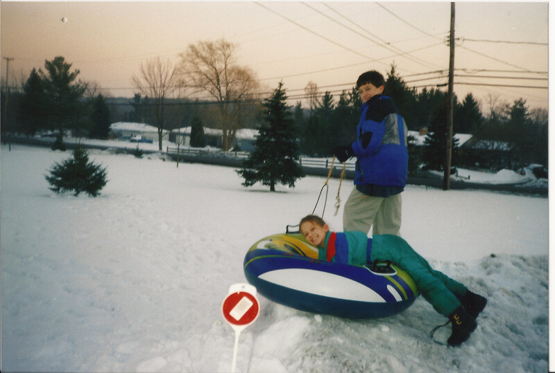 Child sitting in snow tube while adult stands nearby holding rope in winter landscape.
