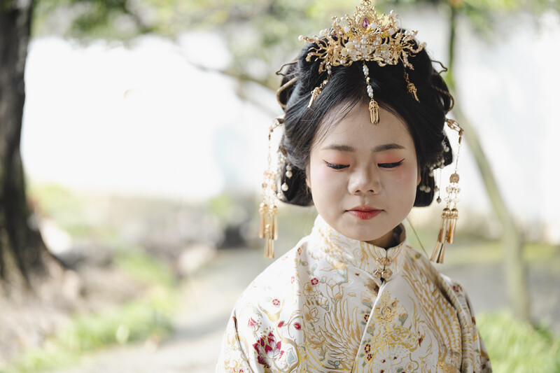 Young child wearing ornate golden headdress and embroidered white garment with downward gaze.