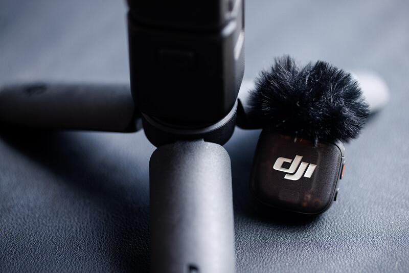 DJI lavalier microphone with windscreen attached next to microphone boom arm on gray surface.
