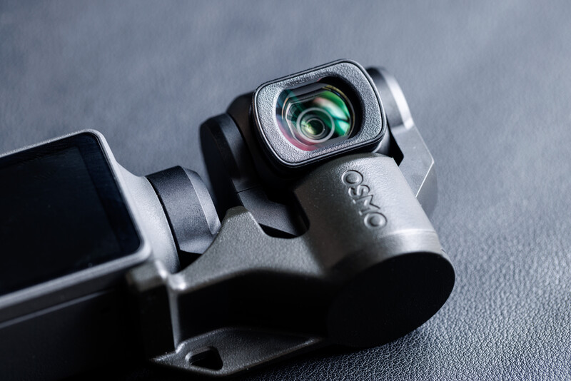 Close-up of a gimbal-mounted camera lens with green optical glass visible against a gray textured surface.