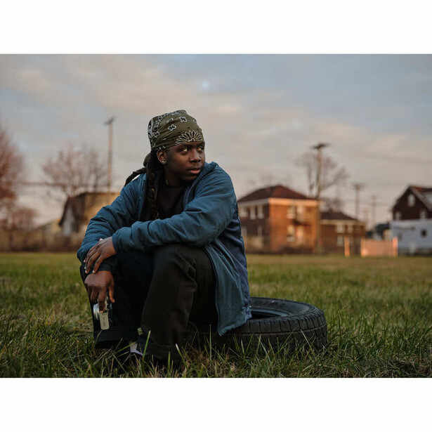 Person in blue jacket and knit beanie squatting on grass with suburban houses and utility poles in background.