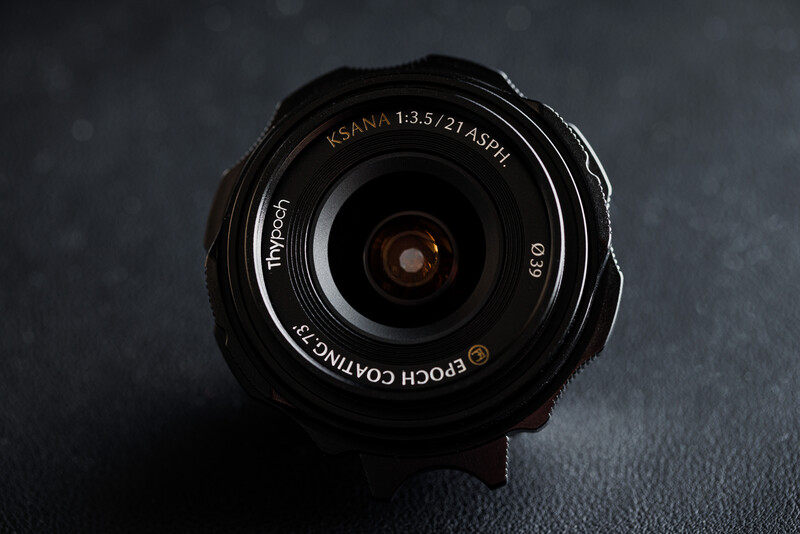 Overhead view of a black KSANA 1:3.5 wide-angle lens on dark textured surface.