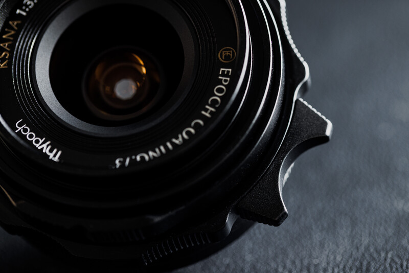 Close-up of a wide-angle camera lens with glass elements visible through the front element, shot against a neutral gray background.