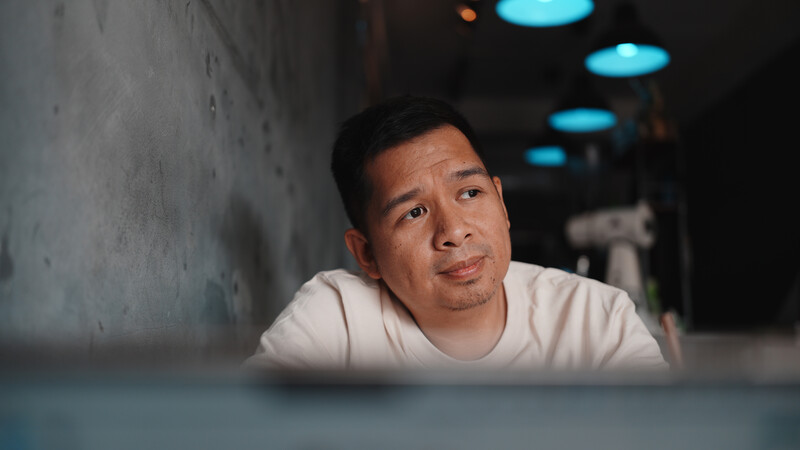Man in white t-shirt leaning against concrete wall in dimly lit corridor with blue overhead lights.