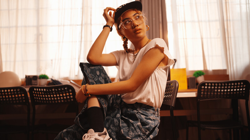 Young woman sitting indoors adjusting sunglasses with a contemplative expression.