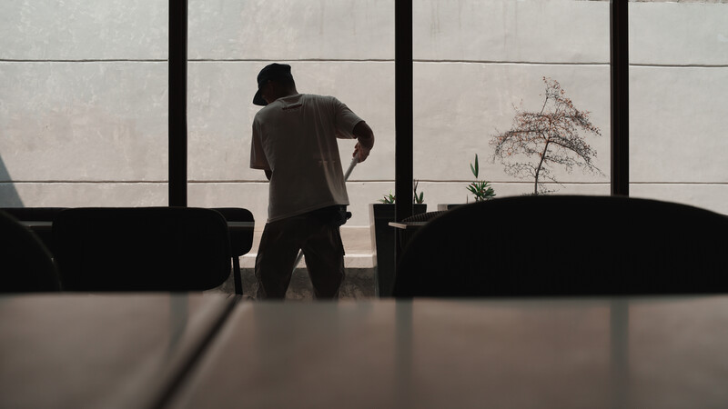 Silhouetted figure standing at large windows overlooking a minimalist interior with plants and furniture.