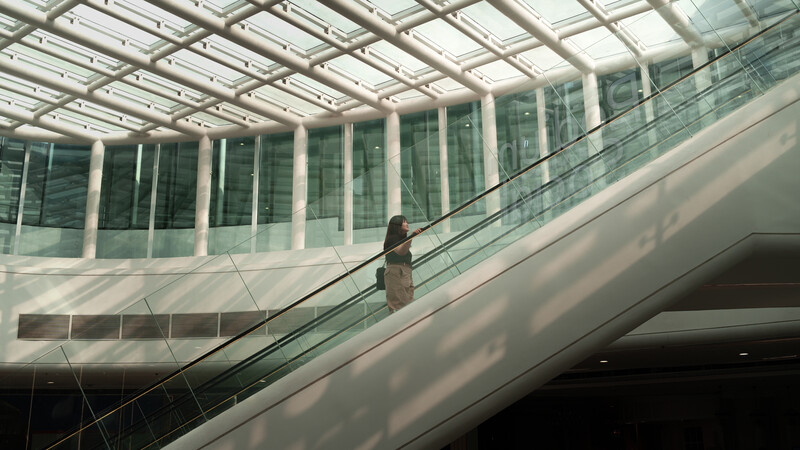 Woman on escalator in modern glass and steel atrium with geometric skylight.