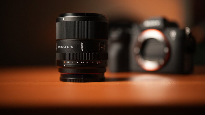Prime lens with focus ring and depth-of-field markings in sharp focus, with camera body and additional lens blurred in background.