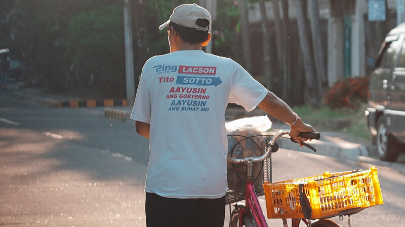Man riding a bicycle with yellow crates attached, wearing a white t-shirt and cap on a sunny street.