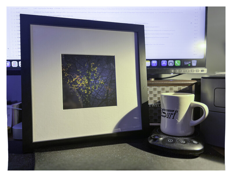 Framed digital display showing abstract golden particles on dark background, positioned on desk next to coffee mug.