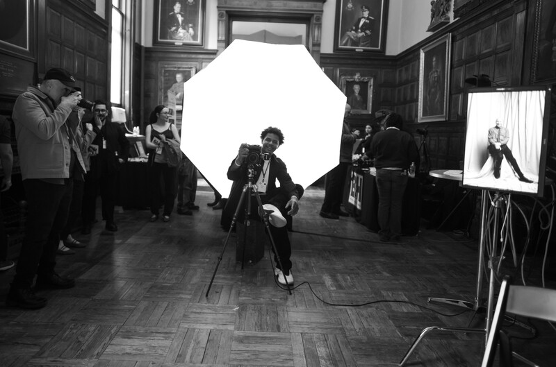 Portrait subject seated under large octagonal softbox during studio photography session in classical interior space.