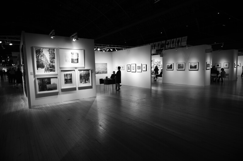 Black and white photograph of a modern gallery interior with white walls displaying framed photographs and artwork.