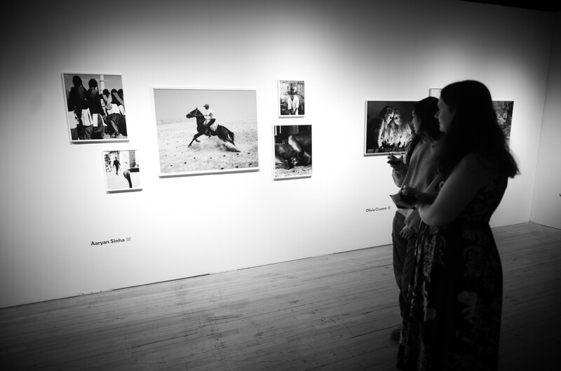 Two visitors viewing a gallery wall of black and white photographs in a minimalist exhibition space.