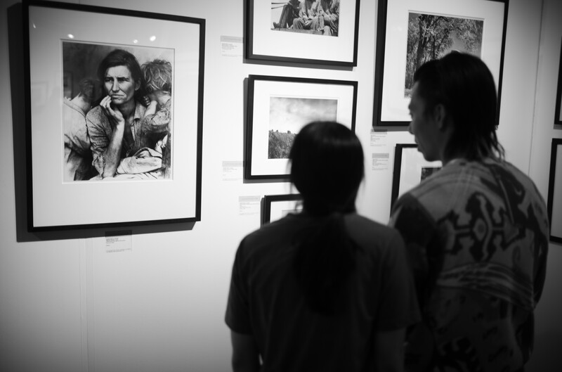 Two visitors view black and white framed photographs displayed on a gallery wall.