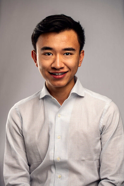 Young man wearing a light gray button-up shirt smiling at camera against neutral gray background.