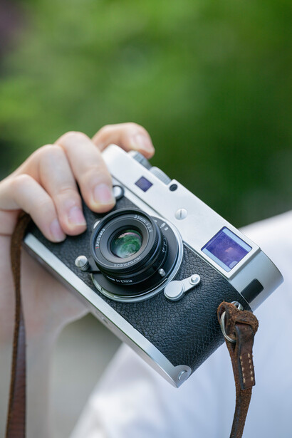 Photographer holding a rangefinder film camera with leather strap against blurred green background.