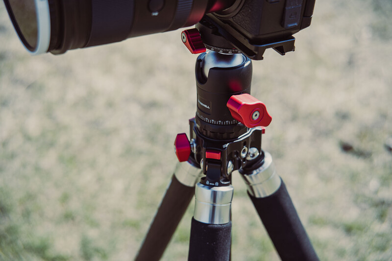 Close-up of a camera mounted on a tripod with a red ball head and black telescoping legs against a blurred outdoor background.