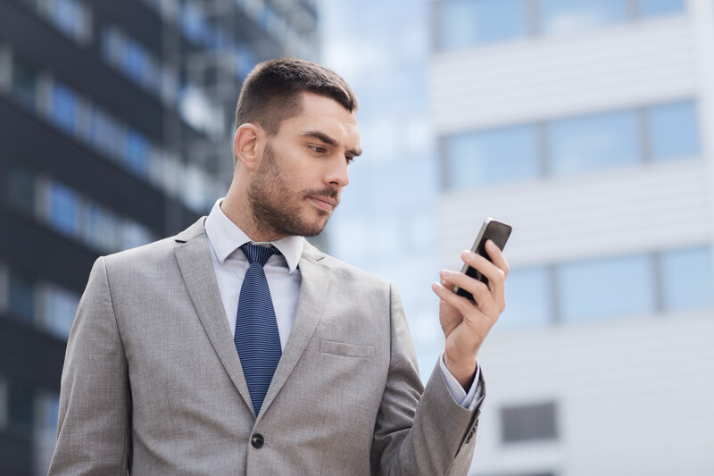 Businessman in gray suit and blue tie holding smartphone outdoors with modern office buildings in background.