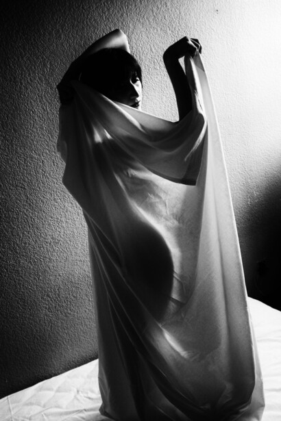 Person wrapped in translucent fabric against white wall with dramatic side lighting creating sharp shadows.