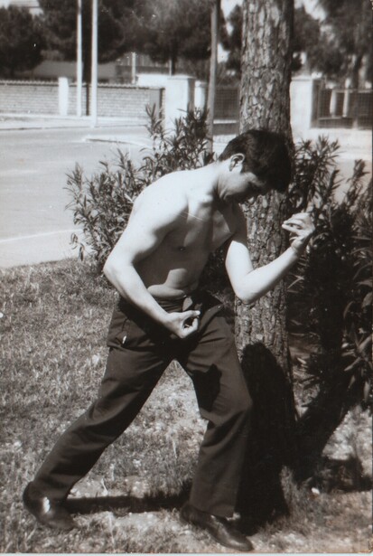 Shirtless man leaning against a tree trunk in an outdoor setting.